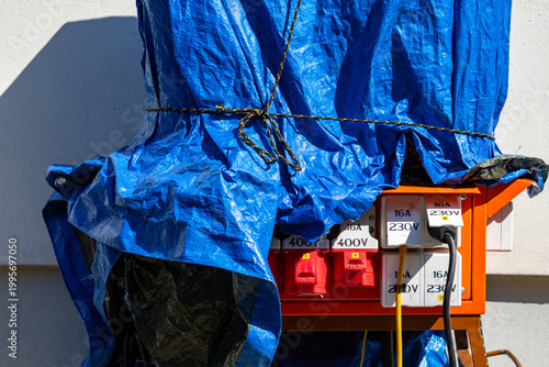 Portable construction power distribution box with 230V and 400V industrial sockets protected by a blue tarp at a work site, temporary electrical installation, professional construction site equipment.