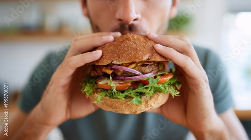 Male holding a large hamburger with lettuce, tomato, and onion, preparing to take a bite in a bright kitchen with blurred background elements