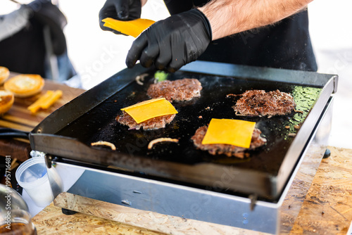 Chef preparing smash burgers on a flat top grill, adding toppings to freshly cooked beef patties with melted cheese, capturing street food cooking process and professional burger preparation in a