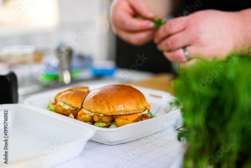 Chef assembling fish burgers, placing toppings on freshly grilled beef patties with melted cheese, showcasing handmade gourmet burger preparation and professional street food cooking process.