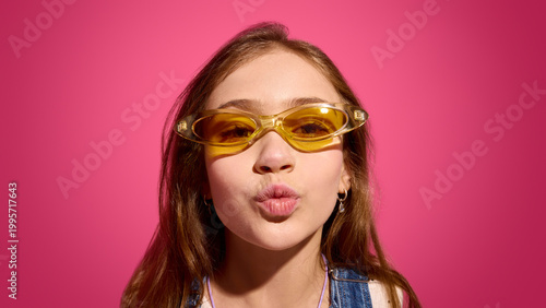 Billede på lærred Child girl in yellow sunglasses blowing kiss on pink background with bright studio lighting and centered composition