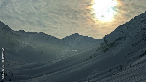 Sunlight breaks through clouds over a snow-covered valley in the Austrian Alps, highlighting the rugged landscape. Ideal for travel documentaries or nature features.