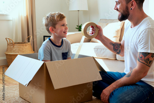 Caucasian father and young son packing cardboard box with tape, smiling during home move and family organization. Ideal for relocation services, parenting, storage, moving day marketing