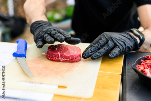 Close-up of a chef in black nitrile gloves precisely trimming a thick-cut ribeye steak with a sharp knife on a cutting board during a professional Steak Cookoff Association (SCA) BBQ competition.