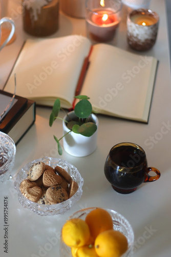 Cup of tea or coffee, teapot, book, notebook, e-reader, glasses, cookies, nuts, lemons, house plants and lit candles on the table. Hygge at home. Selective focus.