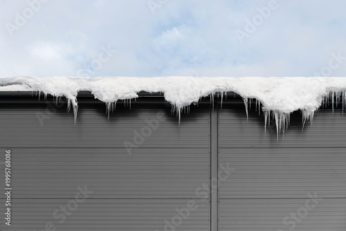 Fotografía Thick layer of white snow and icicles hang from the edge of a grey metal roof against the sky