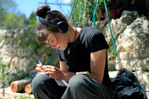 Young woman with bun and headphones sitting outdoors on stone steps, intently writing with a pen in a small notebook, surrounded by lush greenery.