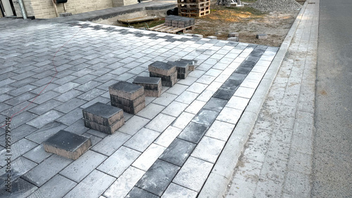 Paved walkway under construction with gray concrete blocks arranged in a pattern, surrounded by unfinished landscaping and construction materials