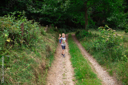 Tableau sur toile Happy little boy waving hand while walking alone on forest path surrounded by lu