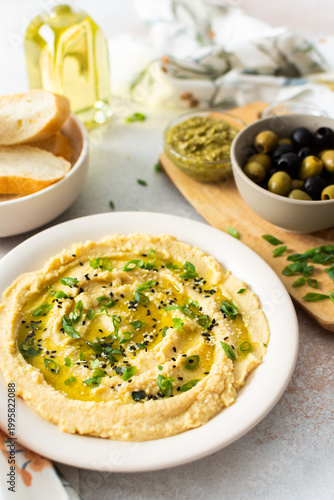 A plate of smooth, creamy hummus drizzled with olive oil sits on a dining table. It is accompanied by bowls of olives, pickles, and fresh bread ready to enjoy