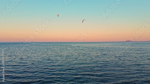 The sky shows warm colors as the sun sets over the ocean. Waves gently move in the water. A distant landmass is visible against the horizon. Birds fly in the air above the scene.