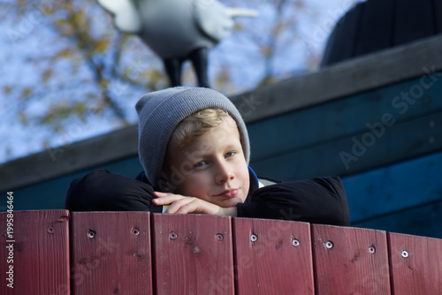 Thoughtful blond boy in grey beanie leaning on a wooden fence