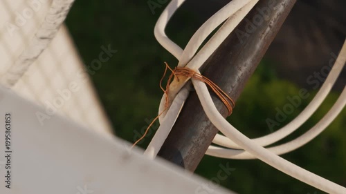 A thick white cable is tied to a rusty metal pole using a thin piece of copper wire. The makeshift connection is outdoors with a soft blurred background of a fence and green foliage.