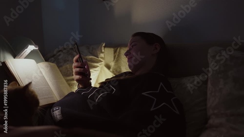 Girl doom scrolling her smartphone instead of reading on a coach at night. Social network and phone addiction concept. Cozy home ambient light