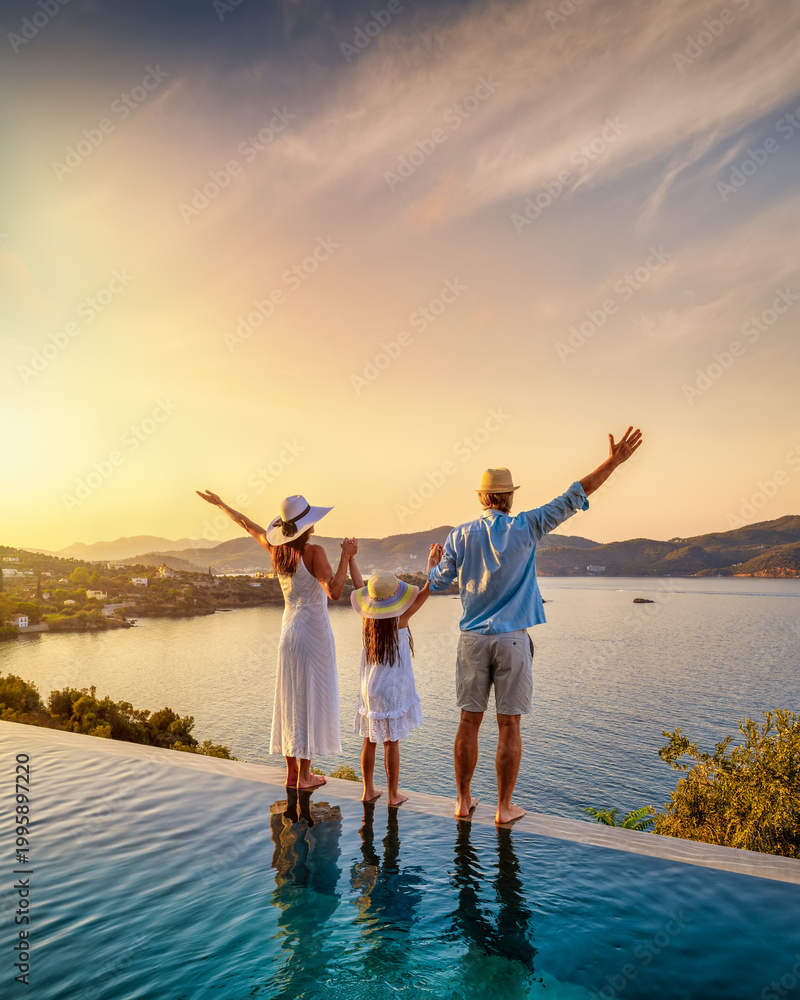 Fototapeta premium A happy family on summer holidays stands by the swimming pool and enjoys the beautiful sunset behind the ocean