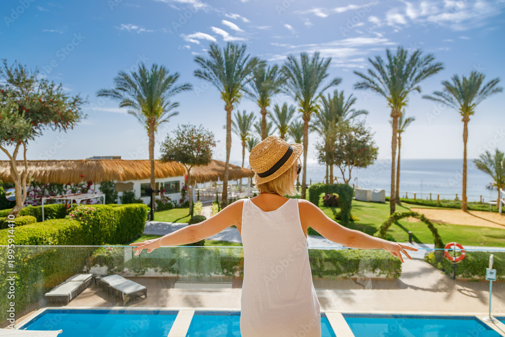 Fototapeta premium Back view of a girl standing on a terrace of a luxury tropical resort, enjoying the ocean view and sunny weather during her summer vacation trip