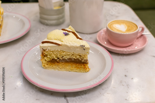 Porción de torta tres leches y vainilla con relleno de dulce de leche y glaseado blanco. Interior minimalista de cafetería con café decorado con forma de corazón. Imagen con desenfoque.