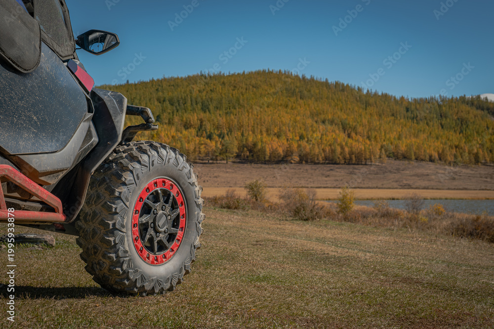 Obraz premium Close-up of off-road buggy UTV wheel with red beadlock parked on a field against a background of autumn larch forest and lake. Extreme adventure trip concept in wild nature under blue sky.