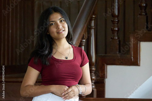 Woman in maroon top standing near wooden staircase neutral expression poised demeanor