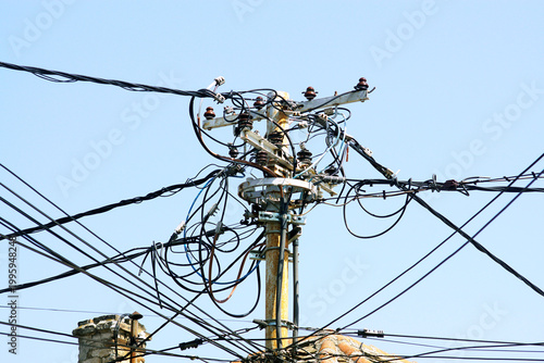 Rusted utility pole crowded with looping cables and ceramic insulators against sky. Captures failing grid infrastructure, electrical distribution chaos, and urgent power network modernization needs.