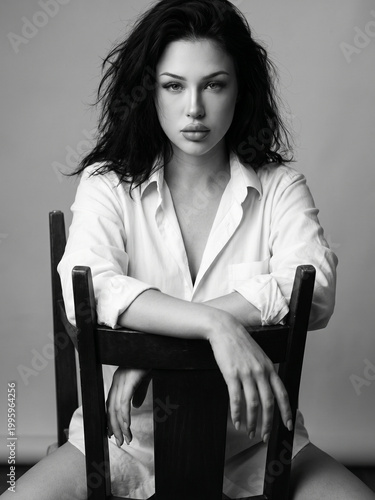 Dramatic black and white portrait of a beautiful young woman with messy bob hair sitting on a wooden chair, moody studio lighting
