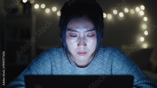 Young Asian woman intensely focused on a laptop screen in a dark room, her face illuminated by the blue light, symbolizing late-night work or study.