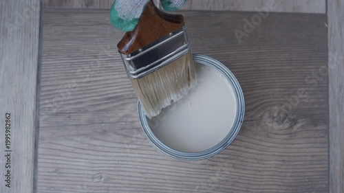 Top view of gloved hand dipping brush into white paint on wood, DIY renovation and surface coating concept.
