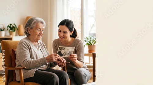 Happy mature granddaughter helping senior grandmother looking at vintage photo album in cozy living room family history memories elderly care nursing home or assisted living