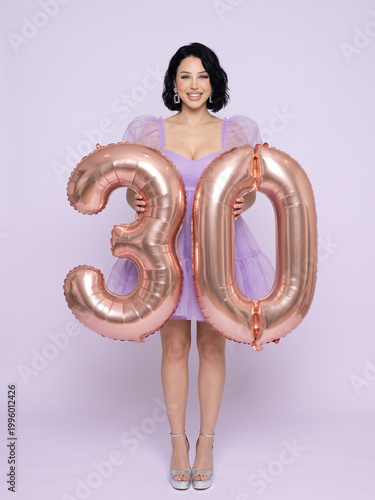 Happy young woman celebrating 30th birthday holding rose gold number balloons, studio portrait on purple background