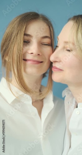 Vertical video of beautiful mature mother kissing her adult daughter wearing in white shirts in the photostudio on blue paper background. Two female generations. Mother's day concept