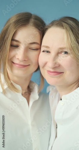 Vertical video of adult daughter in white shirts kissing her beautiful mature mother in the photostudio on blue paper background. Two female generations. Mother's day concept