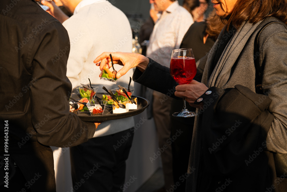 Obraz premium Guests enjoying appetizers and cocktails at a stylish outdoor reception, a server offering canapés as people socialize in warm evening light.
