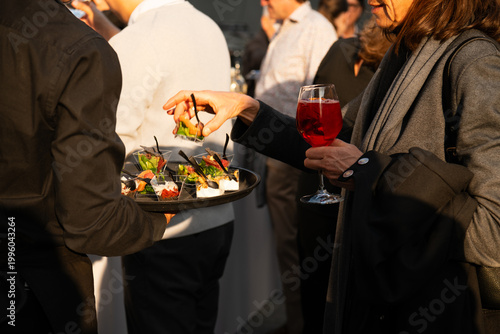 Guests enjoying appetizers and cocktails at a stylish outdoor reception, a server offering canapés as people socialize in warm evening light.