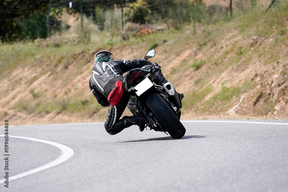 Obraz premium Motociclista con traje de cuero inclinándose en una curva cerrada en carretera de montaña