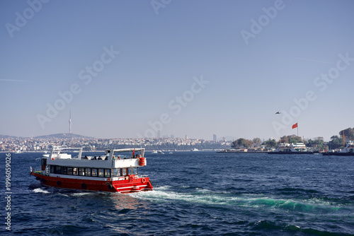 Photography Red passenger boat cruising across the Bosphorus with Istanbul skyline, Turkish flags and clear blue sky on a bright day
