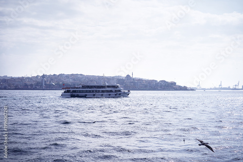 Foto Wide view of a ferry boat navigating through the calm urban waterfront with distant cityscape visible under a cloudy sky, reflecting sunlight and presenting a serene atmosphere