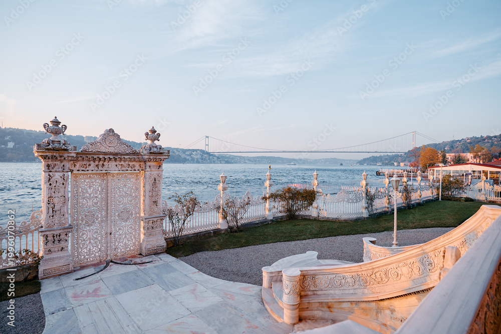 Fototapeta premium Elegan gates frame a waterfront view during sunset, with a bridge spanning the distant horizon and serene surroundings of greenery and architecture creating a picturesque and tranquil landscape.