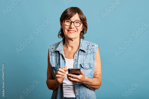 Latina senior woman looking at camera with smartphone on blue background