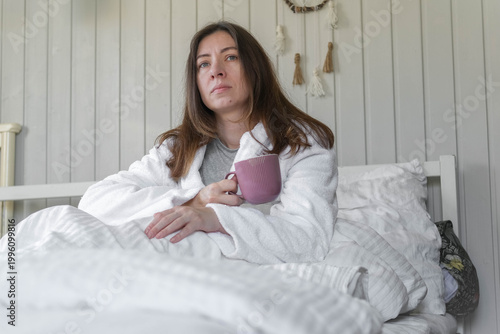 A woman lies on a pastel-colored bed, holding a coffee mug. A calm woman in a robe enjoys the serene morning atmosphere at home. Morning routine. Self-care. Starting a new day. Space for text. Banner