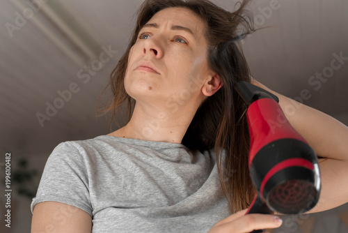 A woman blow-drying her hair, soft morning sunlight shining through the curtains, a concentrated expression on her face, hands running through chestnut curls, a home routine, relaxed self-care.