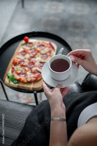 Cropped view of a woman holding a cup of tea while meat pizza on a wooden board stands blurred in the background on a small black table