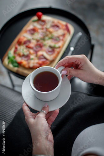 Cropped female hands holding a cup of tea while fresh pizza on a wooden board stands blurred in the background indoors