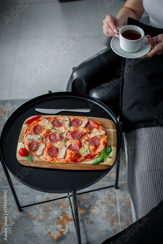 Cropped view of a woman holding a cup of tea near a fresh baked meat pizza served on a wooden board over a small black table