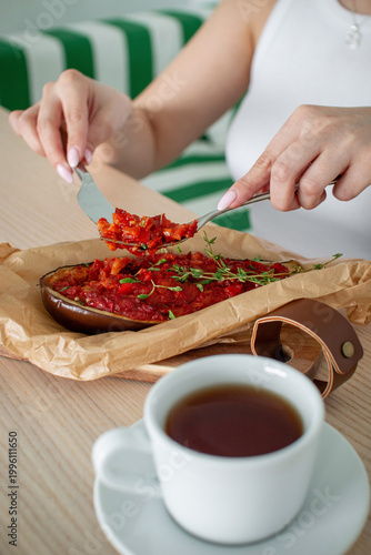 Woman eating baked stuffed eggplant with rich tomato sauce and herbs, served on wooden board with hot tea in cozy cafe setting