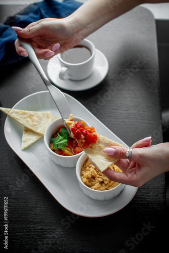 Female hands making a flatbread snack with hummus and roasted vegetable dip from a white appetizer platter with tea on dark table