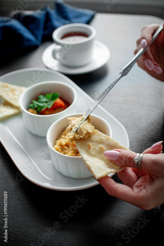Female hands spreading hummus on flatbread from a white appetizer platter with roasted vegetable dip and tea on dark restaurant table