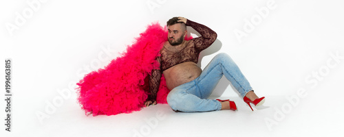 Horizontal photo. Mature man with beard, big belly, pink magenta tulle fur coat, mesh through tank top, blue denim jeans, red stiletto high heels sits on floor on white. LGBT pride month concept