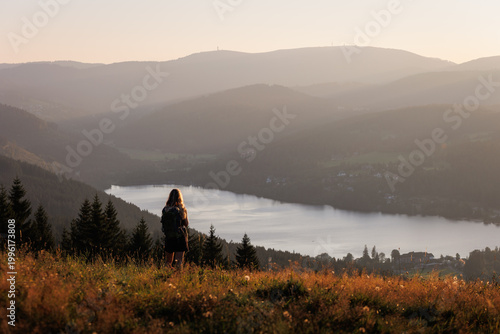 Goldene Abendsonne taucht den Titisee im Schwarzwald in warmes Licht, während eine Person auf einer Wiese steht und über das Tal, den See und die bewaldeten Hügel blickt.