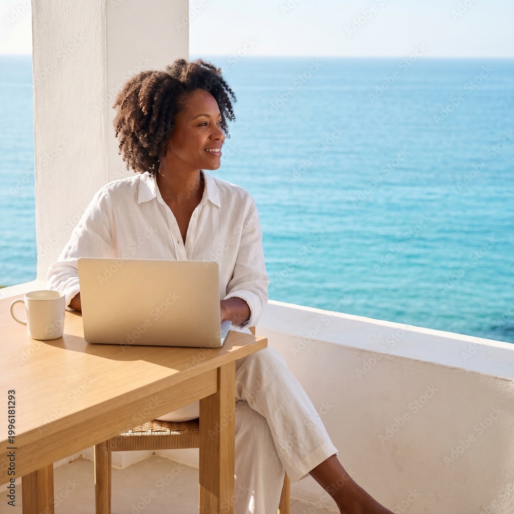 Fototapeta premium A happy African American woman works remotely on her laptop from a sunny balcony overlooking the beautiful blue ocean, enjoying the freedom of a digital nomad lifestyle in a luxury resort