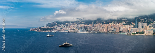 Tableau sur toile A wide aerial panorama shows Monaco on the French Riviera, with high rises and terraced blocks around a deep bay as yachts cruise and anchor under low clouded hills